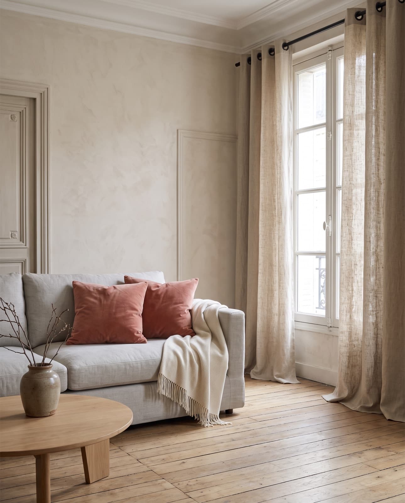 Un salon clair : canapé gris aux coussins argile et au plaid écru, fenêtre aux rideaux en lin naturel