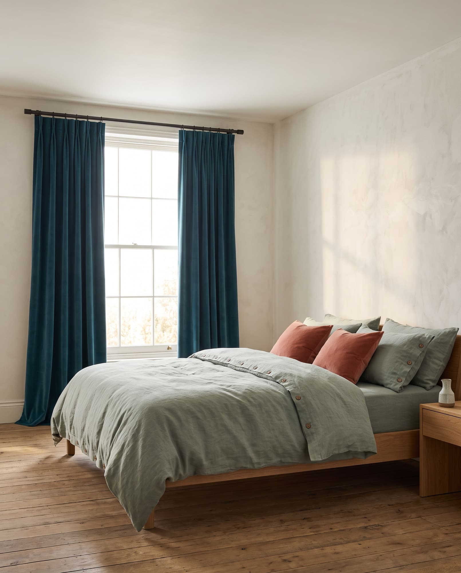 Une chambre au petit matin : lit en lin vert sauge, coussins en velours argile et hauts rideaux de velours bleu canard